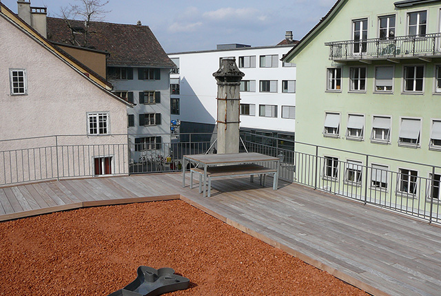 Ein Ausschnitt der Dachterrasse, mit Holzbodenplatten und in der Mitte roter Sand. Eine Sitzgelegenheit.
