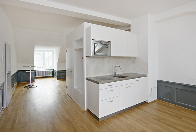 Dachwohnung: ein großzügiger heller Raum mit Garderobe und Küchenzeile, alles in weiß gehalten. Nur die Wände haben im unteren Bereiche eine Holzverkleidung in Grau. Fußboden Holzdielen.