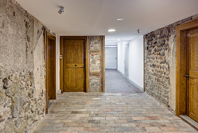 Ein Korridor mit altem Steinfussboden, Holztüren und sehr altem Wandmauerwerk.